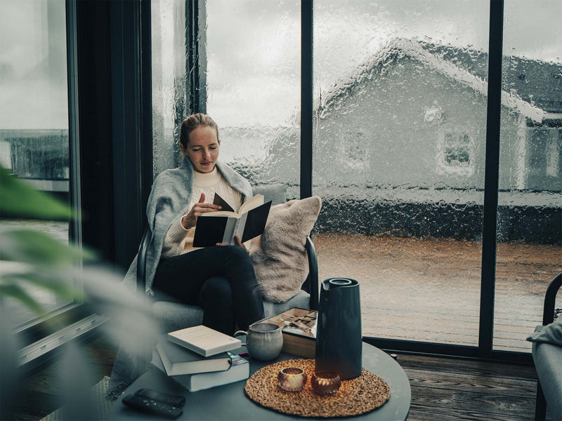 Person liest ein Buch in der Ecke des Wintergartens. Es regnet stark gegen das Schiebeglas. Teelichter auf dem Tisch schaffen eine gemütliche Atmosphäre.